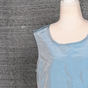 Pu&ograve; includere: Un top senza maniche, azzurro chiaro, realizzato in materiale serico. Il top ha uno scollo tondo ed &egrave; esposto su un manichino. Lo sfondo &egrave; un tessuto grigio strutturato.