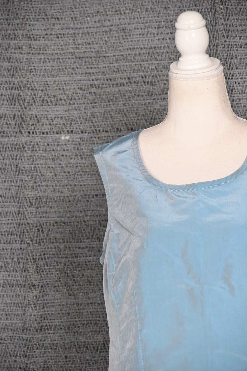Pu&ograve; includere: Un top senza maniche, azzurro chiaro, realizzato in materiale serico. Il top ha uno scollo tondo ed &egrave; esposto su un manichino. Lo sfondo &egrave; un tessuto grigio strutturato.