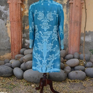 May include: A teal long coat with a floral design, presented on a wooden mannequin. The coat has a high collar and long sleeves. The floral pattern is in a lighter teal shade. The coat is displayed outdoors, with a stone backdrop.