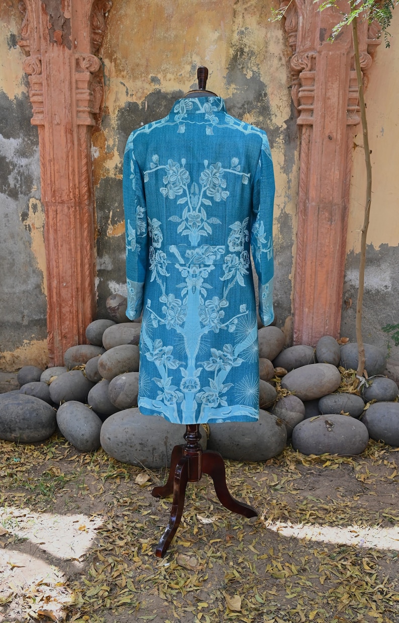 May include: A teal long coat with a floral design, presented on a wooden mannequin. The coat has a high collar and long sleeves. The floral pattern is in a lighter teal shade. The coat is displayed outdoors, with a stone backdrop.