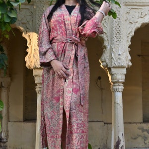 May include: A long, flowing robe with a red and beige paisley pattern. The robe is belted at the waist and has long sleeves. The person is wearing black sunglasses and black sandals.