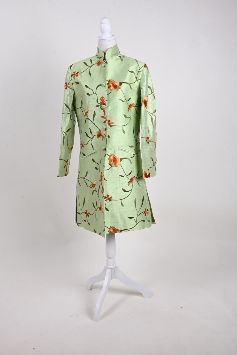 May include: A light green, long-sleeved coat with a mandarin collar and floral embroidery. The coat is displayed on a white mannequin. The floral design features orange and brown flowers and vines.
