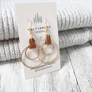 以下が含まれることがあります： 茶色の革のアクセントが付いたゴールドのフープイヤリング。イヤリングは、"Salt + Shore Designs"というテキストとウェブサイトアドレスが書かれた白いカードの上にあります。
