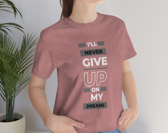 May include: A heather pink t-shirt with a white graphic that reads "I'll Never Give Up On My Dreams". The text is in a bold, blocky font.