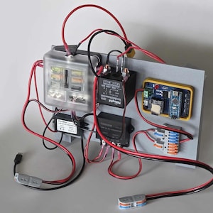 May include: A circuit board with various electronic components, including a clear fuse box, black relays, and a yellow-framed microcontroller. Red and black wires connect the components.