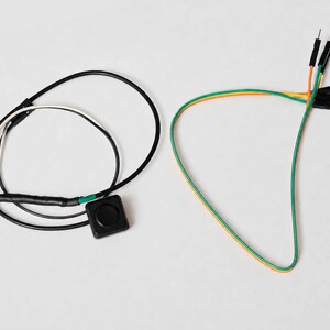 Pode incluir: Uma vista a&eacute;rea mostra dois componentes eletr&ocirc;nicos contra um fundo branco. Um &eacute; um bot&atilde;o preto com fios, e o outro &eacute; um conjunto de fios coloridos com pinos. Os fios s&atilde;o verdes, laranja e amarelos.