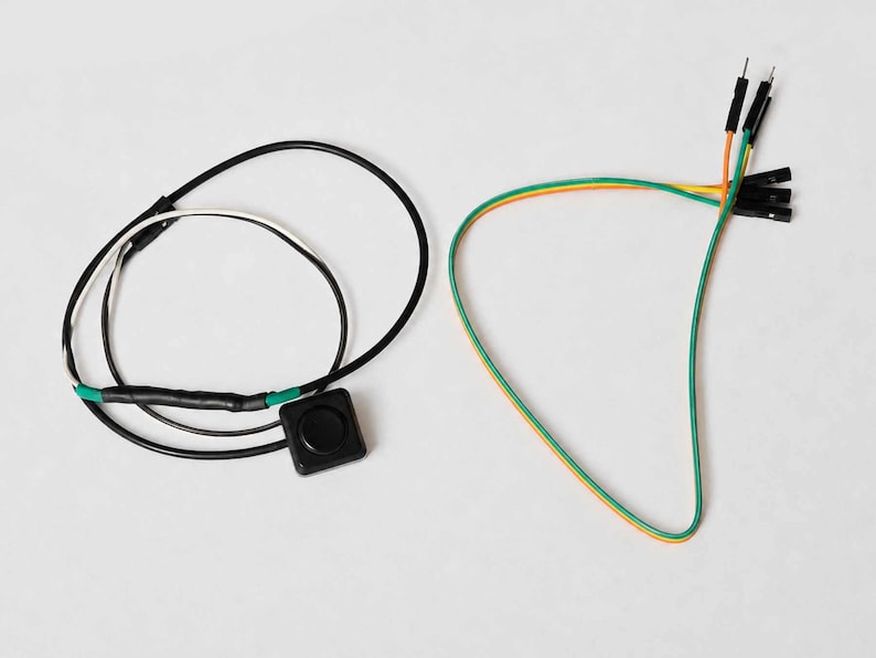 Pode incluir: Uma vista a&eacute;rea mostra dois componentes eletr&ocirc;nicos contra um fundo branco. Um &eacute; um bot&atilde;o preto com fios, e o outro &eacute; um conjunto de fios coloridos com pinos. Os fios s&atilde;o verdes, laranja e amarelos.