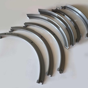May include: A collection of curved, metallic machine parts arranged on a white surface. The parts are a dark gray color with a lighter gray accent on the inner edge. Each piece has a unique shape and design, suggesting they are components of a larger mechanical system.