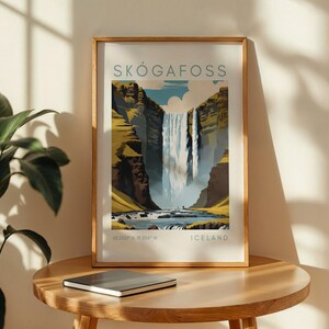 May include: Framed art print of Sk&oacute;gafoss waterfall in Iceland. The artwork features a vibrant illustration of the waterfall cascading down a cliff, with text that reads "SK&Oacute;GAFOSS" and "ICELAND". The print is displayed on a wooden table.