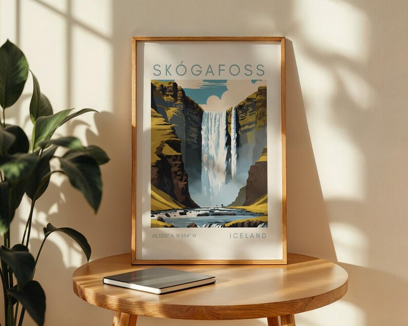 May include: Framed art print of Sk&oacute;gafoss waterfall in Iceland. The artwork features a vibrant illustration of the waterfall cascading down a cliff, with text that reads "SK&Oacute;GAFOSS" and "ICELAND". The print is displayed on a wooden table.