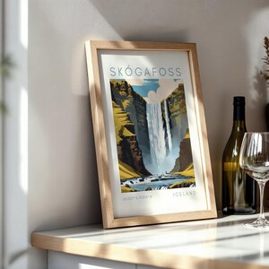 May include: Framed art print of Sk&oacute;gafoss waterfall in Iceland. The artwork features a vibrant depiction of the waterfall cascading down a cliff, with the text "SK&Oacute;GAFOSS" at the top and "ICELAND" at the bottom. The frame is made of light-colored wood.