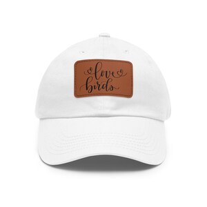 Puede incluir: Gorra de béisbol blanca con un parche de cuero marrón. El parche presenta las palabras "love birds" en fuente cursiva. La gorra tiene una visera curva y un diseño clásico de seis paneles.