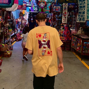 May include: A person wearing a yellow t-shirt with a graphic of a playing card featuring a skeleton with wings and hearts. The card is the Ace of Hearts. The person is walking through a market with colorful decorations and crafts.
