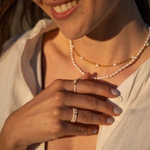 Può includere: Una donna indossa una camicia bianca con una collana di perle a strati e due anelli di perle al dito.