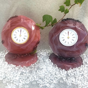 Può includere: Due orologi decorativi con un design sfaccettato e geometrico. Gli orologi sono realizzati in resina viola e rosa con accenti neri. Gli orologi sono su una superficie bianca.