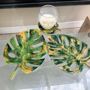 Peut inclure: Trois sous-verres en résine verte et jaune en forme de feuilles de monstera. Les sous-verres sont sur une table en verre avec un verre de lait en arrière-plan.