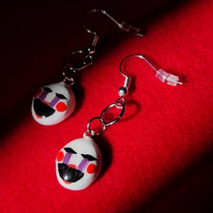May include: A pair of handmade earrings featuring a white, oval-shaped face with black, purple, and red details. The earrings have silver-colored hooks and chain links, set against a red background.