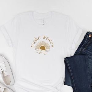 May include: White t-shirt with a gold graphic of a sun setting over waves and the text "make waves". The shirt is paired with blue jeans and white sneakers.