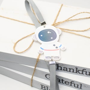 May include: A stack of three white wooden blocks with the words thankful, grateful, and blessed printed on the sides. A gray elastic band wraps around the blocks, featuring a white astronaut charm with blue and pink accents and star details.
