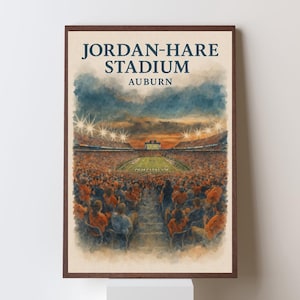 May include: A framed watercolor-style art print depicting Jordan-Hare Stadium in Auburn. The image shows a crowded stadium with orange-clad spectators, a green football field, and bright stadium lights under a dark, moody, cloudy sky.