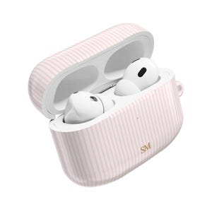 May include: A pink and white striped AirPods case with white AirPods inside. The case is open, showing the earbuds. The initials "SM" are printed in gold on the case.