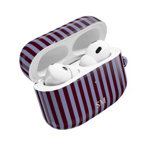 Puede incluir: Una funda abierta para AirPods a rayas burdeos y azul claro, con dos AirPods blancos en su interior. La funda tiene un patrón de rayas a juego y las iniciales "SM" son visibles.