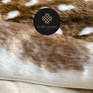 May include: Close-up of a brown and white faux fur rug with a stitched edge. The rug has a pattern of white spots on a brown background. A black circle with a gold logo that says "LUXE LIVING LOUNGE" is visible in the upper right corner of the image.