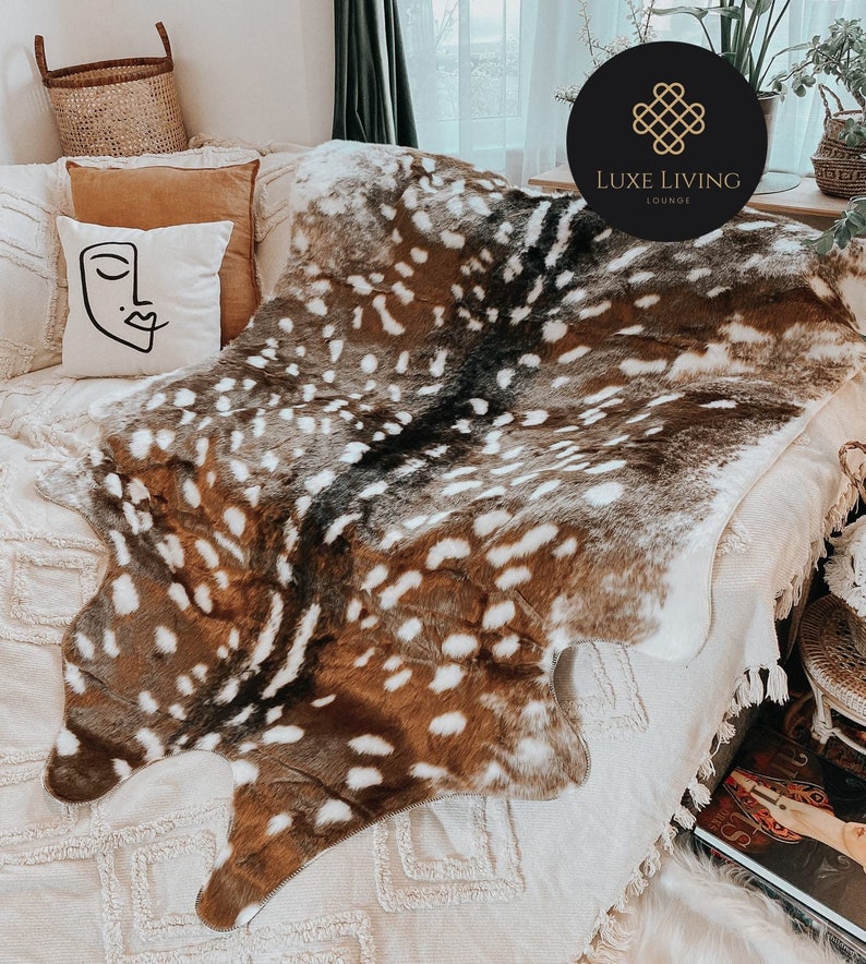May include: A brown and white faux fur throw blanket with a spotted pattern, resembling a deer hide. The blanket is draped over a white couch with a brown pillow and a white pillow with a black abstract face design. The text "LUXE LIVING LOUNGE" is visible in a black circle with a gold border.