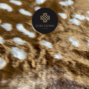 May include: Close-up of a brown and white spotted faux fur fabric. The fabric has a soft, plush texture. A black circle with a white logo that says "LUXE LIVING LOUNGE" is in the upper right corner of the image.