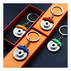 Peut inclure: Quatre porte-clés de rotor de frein de couleurs différentes avec des anneaux de clés argentés. Les porte-clés sont dans des boîtes avec des couvercles orange. Les rotors de frein sont argentés avec des étriers de frein rouge, bleu, vert et jaune.