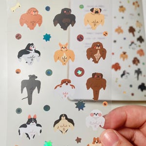 May include: A sheet of clear stickers featuring various breeds of dogs with muscular bodies. The stickers are on a white background with glitter accents.