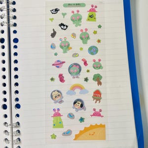 May include: A clear sticker sheet with various cartoon alien and space-themed designs. The stickers include green aliens, planets, rainbows, and the text "All x OoL". The stickers are on a lined notebook page.