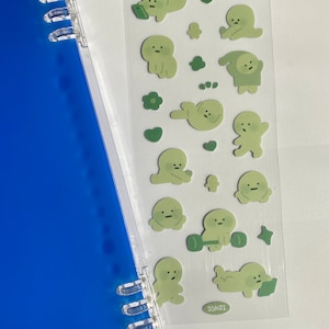 以下が含まれることがあります： 緑色の漫画のキャラクターが描かれた透明なステッカーシート。キャラクターは電話をかけたり、運動したり、寝たりなど、さまざまなポーズをとっています。ステッカーには「SSK01」とラベルが貼られています。