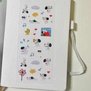 May include: A white open notebook with a dotted grid pattern, featuring a strip of Snoopy-themed stickers. The stickers include Snoopy in various poses, along with musical notes, hearts, clouds, and suns. The notebook has a ribbon bookmark.