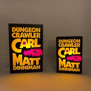 May include: Two illuminated black rectangular signs with the text "Dungeon Crawler" and the names "Carl" and "Matt Dinniman" in yellow. A pink graphic of a figure is also visible. One sign is larger than the other.