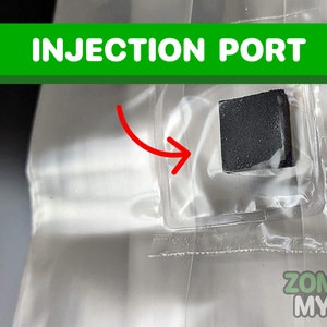 May include: A close-up of a black injection port on a clear plastic bag. The bag is labeled "Zombie Myco".