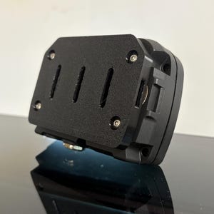 May include: Black rectangular device with a textured surface, featuring three vertical slots and several screws. The device is angled, revealing a side with a small metal component. The object is on a reflective surface.