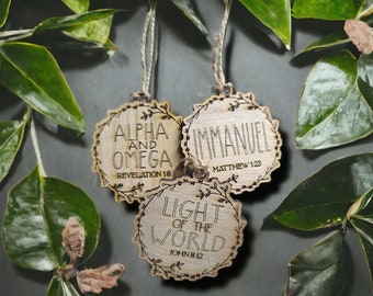 May include: Three wooden ornaments with a floral design and a white background. The ornaments feature the words "Alpha and Omega", "Immanuel", and "Light of the World" with corresponding Bible verses.