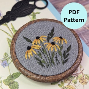 May include: A wooden embroidery hoop with a gray linen fabric background featuring an embroidered design of yellow and black wildflowers. The text "PDF Pattern" is visible in a blue circle.
