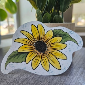 May include: A yellow sunflower with green leaves on a white background with blue watercolor splatters. The sunflower is drawn in a cartoon style.