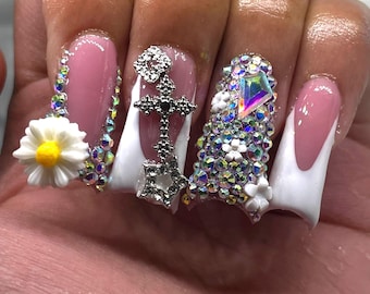 May include: A set of long, embellished artificial nails. The nails feature a pink and white French tip design, adorned with rhinestones, floral accents, and cross pendants. The nails are decorated with a variety of iridescent gems and white floral accents.