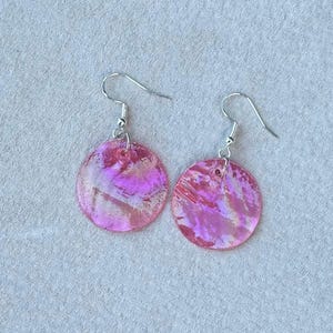 May include: A pair of iridescent pink disc earrings with silver-colored hooks. The earrings have a marbled effect, with shades of pink and white. The earrings are round and appear lightweight.