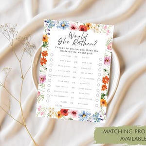 May include: A rectangular game card titled Would She Rather with a floral border of colorful wildflowers. The text includes a list of choices for a bride-to-be, such as stay home or go out, own a dog or a cat, drink coffee or tea, and shop locally or online.