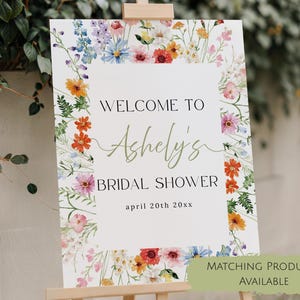 May include: A rectangular sign with a white background and a colorful watercolor floral border. The text reads WELCOME TO Ashely's BRIDAL SHOWER april 20th 20xx. The sign is displayed on a wooden easel stand, featuring various wildflowers in shades of orange, pink, blue, and green.