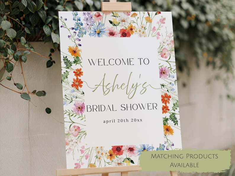 May include: A rectangular sign with a white background and a colorful watercolor floral border. The text reads WELCOME TO Ashely's BRIDAL SHOWER april 20th 20xx. The sign is displayed on a wooden easel stand, featuring various wildflowers in shades of orange, pink, blue, and green.