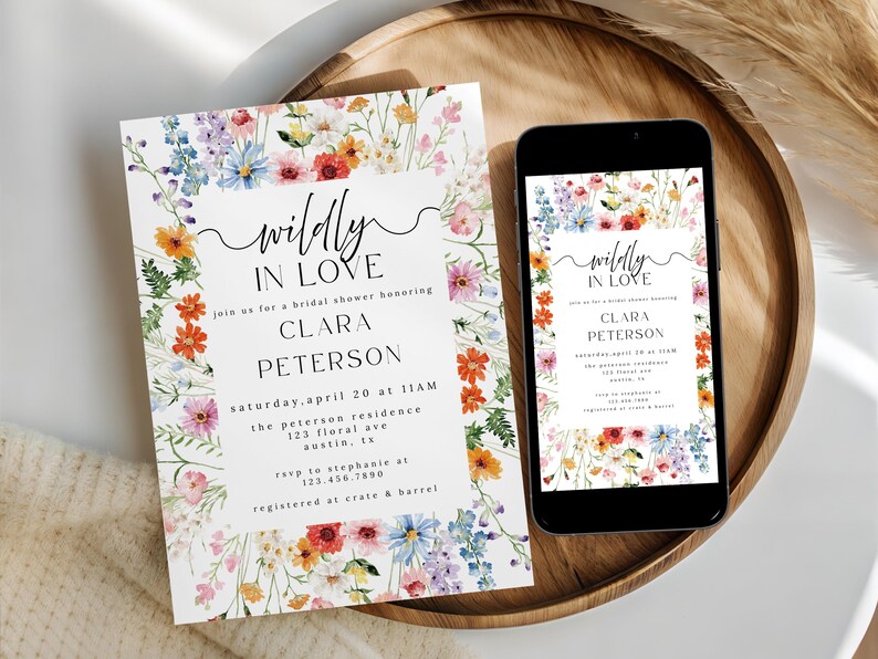 May include: A rectangular bridal shower invitation and a smartphone displaying the same design, both featuring a colorful watercolor wildflower border and black text that reads "wildly IN LOVE" followed by event details for Clara Peterson.