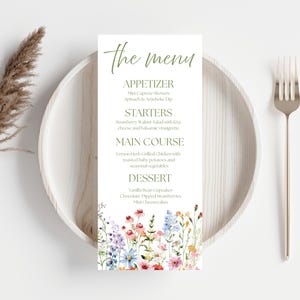 Può includere: Un biglietto del menu con il testo "the menu" in un carattere corsivo. Il menu elenca antipasti, antipasti, piatti principali e dessert. La parte inferiore della carta presenta un motivo floreale ad acquerello. Un piatto di legno, forchetta e coltello sono a destra.