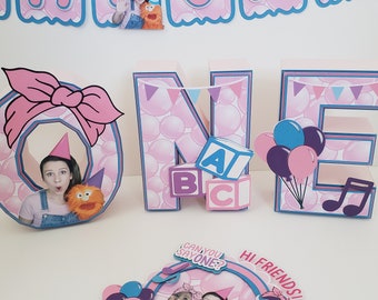 Könnte beinhalten: Ein rosa-blaues 'ONE' Geburtstagsdekorationsset. Das 'O' hat eine rosa Schleife und ein Foto einer Person. Das 'N' hat ABC-Blöcke, und das 'E' hat Luftballons und Noten. Ein Banner mit dem Wort 'ONE' befindet sich im Hintergrund.