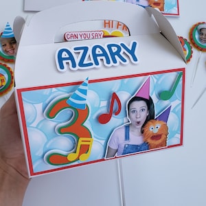 Puede incluir: Cajas blancas para regalos de fiesta con un diseño azul y rojo. Las cajas presentan el nombre "Azary" y el número "3" con notas musicales. Las cajas tienen una foto de una mujer y una marioneta. También se ven adornos adicionales para cupcakes.
