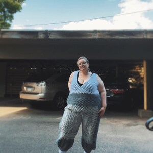 May include: A person wearing a light blue crocheted jumpsuit with a black bra peeking out. The jumpsuit has a textured, puffy design. The person is standing in a parking lot with a car in the background.